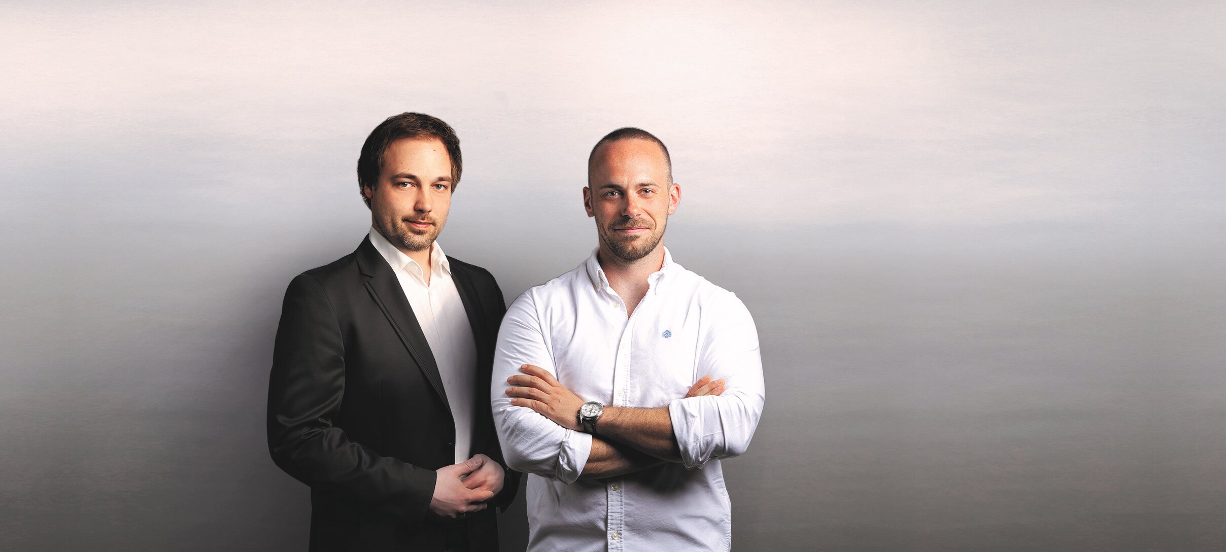 Zwei Männer stehen vor weißer wand, schick angezogen, gucken in Kamera und lächeln | © ecozins